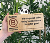 Hand holding a B Corp wooden plaque with The Cheeky Panda's logo peaking behind the plaque.